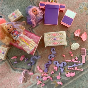 May include: A collection of vintage doll furniture and accessories in shades of pink, purple, and white. Includes a doll, bed, dresser, mirror, and various decorative items like seashells and hair accessories. The doll has long blonde hair.