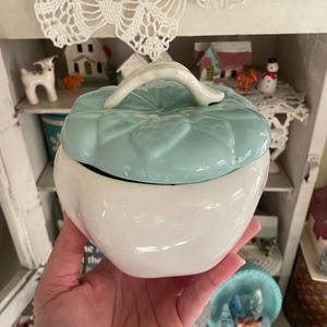 May include: A two-toned ceramic container with a lid. The base is white, and the lid is light blue with a textured, quilted design and a white handle. The container is held in a hand, with other decorative items visible in the background.