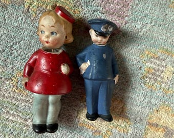 Pode incluir: Duas figuras de cerâmica vintage. Uma é uma boneca com um vestido vermelho e um chapéu vermelho. A outra é um policial com uniforme e chapéu azul. Ambas as figuras têm rostos pintados e estão em pé.
