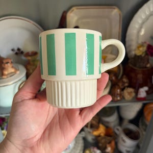 May include: A cream-colored ceramic mug featuring vertical green stripes on the upper portion and a ribbed, fluted texture on the lower base. The mug includes a curved handle on the right side and has a smooth, glossy finish throughout the exterior.