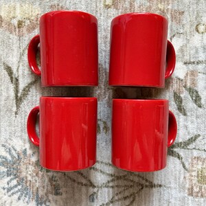 May include: A set of four identical, cylindrical ceramic mugs in a vibrant, solid red color. Each mug features a smooth, glossy finish and a curved handle on the side. The four mugs are arranged in a two-by-two grid formation, showing their exterior.