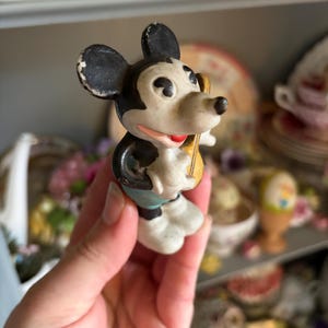 May include: A vintage Mickey Mouse figurine, featuring black ears, a white face, and a red mouth. The figure is holding a yellow object and wearing blue shorts and white gloves. The figurine is a collectible item.