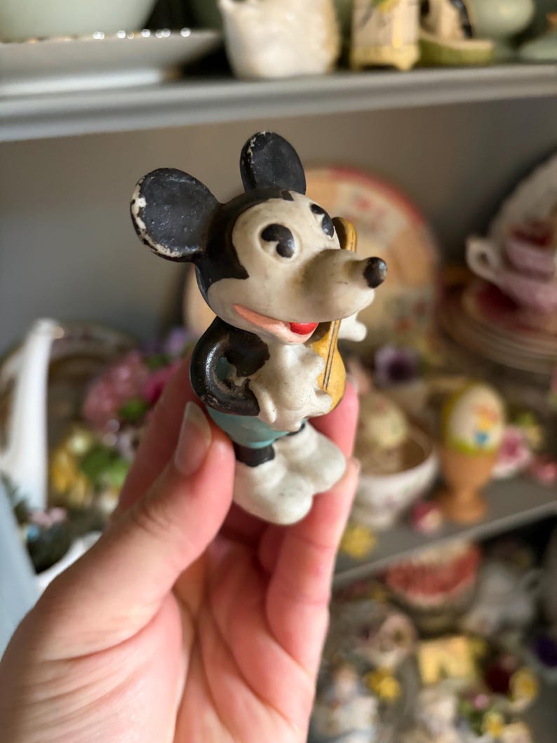 May include: A vintage Mickey Mouse figurine, featuring black ears, a white face, and a red mouth. The figure is holding a yellow object and wearing blue shorts and white gloves. The figurine is a collectible item.