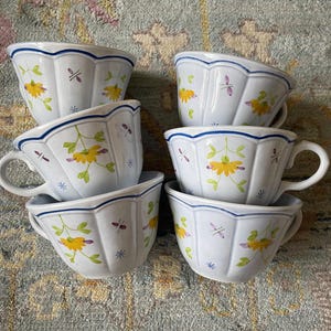 May include: Six white ceramic teacups with scalloped edges and handles. Each cup features a yellow floral design with green leaves and purple accents. The rims are trimmed with a blue line.