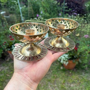 May include: Two vintage brass candle holders. Each has a round base with a decorative cut-out design, a pedestal, and a bowl-shaped top with a floral pattern. The candle holders are held in a hand against a blurred green background.