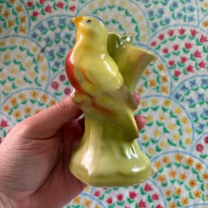 May include: A yellow ceramic bird figurine with a red breast and blue head, perched on a green tree branch. The bird has a small opening on its back, possibly for holding small items. The figurine is hand-painted.