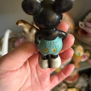 May include: Vintage Mickey Mouse figurine, black with large ears, wearing blue shorts with yellow buttons. The text "MICKEY MOUSE" is visible on the shorts. The figurine has white gloves and shoes. A collectible Disney character.