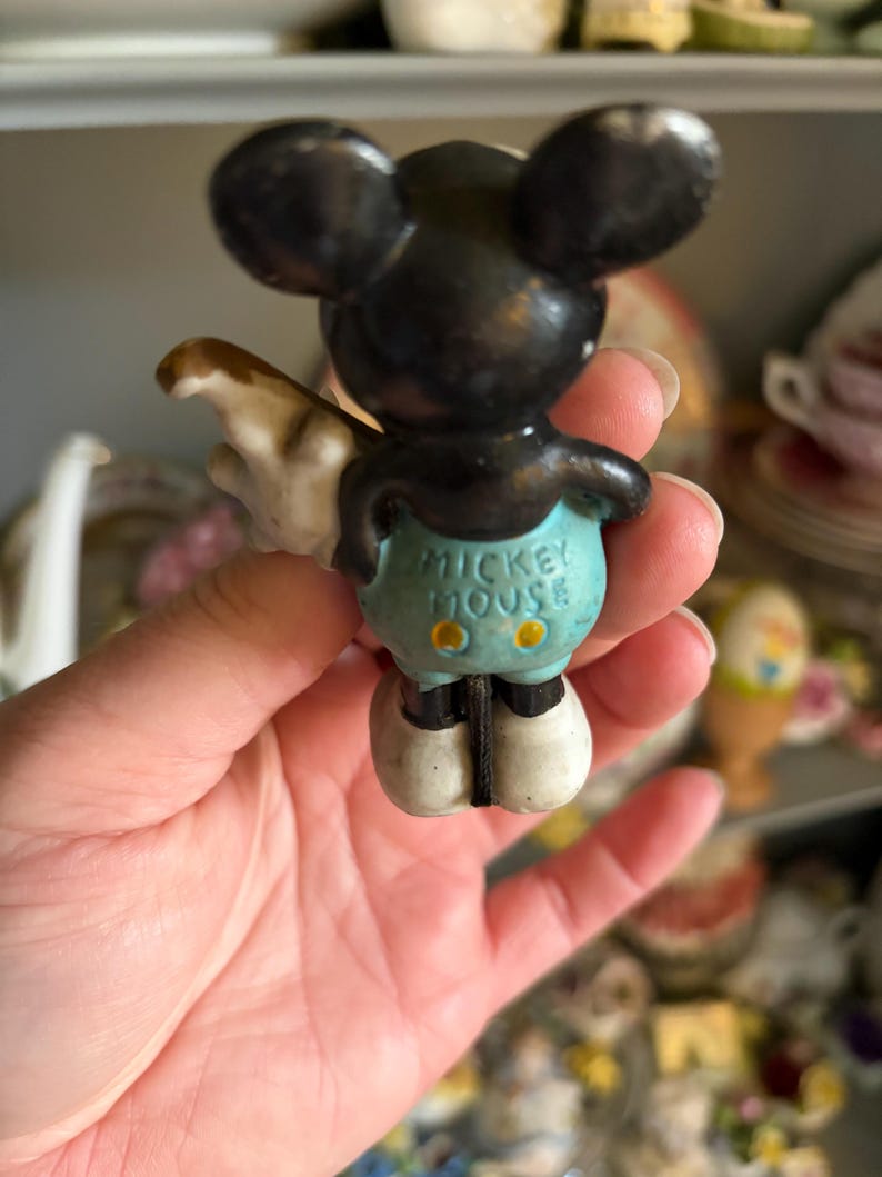 May include: Vintage Mickey Mouse figurine, black with large ears, wearing blue shorts with yellow buttons. The text "MICKEY MOUSE" is visible on the shorts. The figurine has white gloves and shoes. A collectible Disney character.
