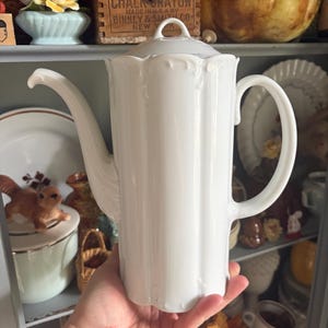 May include: A tall white porcelain coffee pot featuring a vertical fluted design, a curved handle, a long slender spout, and a matching lid with a small circular knob handle. The body has a scalloped rim and a smooth, glossy finish.