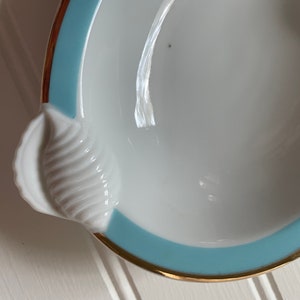 May include: White seashell-shaped dish with a light blue border and gold trim. The dish has a detailed, textured seashell design on one side. A decorative piece for serving or display.