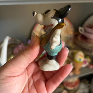 May include: A vintage Mickey Mouse figurine, featuring black ears, a white face, and blue shorts. The figurine is holding a small, brown instrument. The figure is a collectible item.