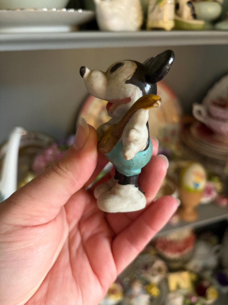 May include: A vintage Mickey Mouse figurine, featuring black ears, a white face, and blue shorts. The figurine is holding a small, brown instrument. The figure is a collectible item.