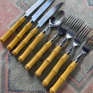 May include: A set of stainless steel flatware with bamboo-style yellow handles. The set includes knives, spoons, and forks. The utensils are arranged on a patterned surface, suggesting a dining or serving context.