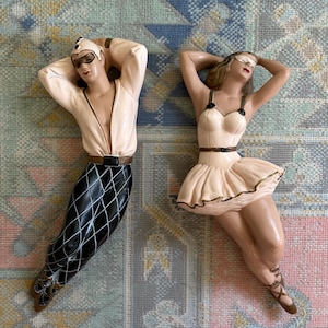 May include: Two decorative figurines, one male and one female, in a relaxed pose. The male figure wears a white top, black and white diamond-patterned pants, and sunglasses. The female figure is in a cream-colored outfit with a skirt and a mask.