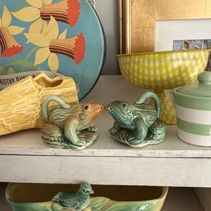 May include: A pair of ceramic frog figurines in shades of green and orange, perched on a shelf. The frogs have detailed features and are positioned on lily pad bases. Other items include a yellow and white gingham bowl, a blue floral tray, and a green and white striped jar.