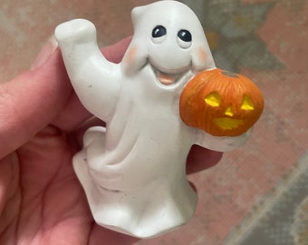 May include: A white ceramic ghost figurine with a friendly expression, rosy cheeks, and black eyes. The ghost is waving one hand while holding a small orange jack-o'-lantern pumpkin with a carved yellow face in the other hand.