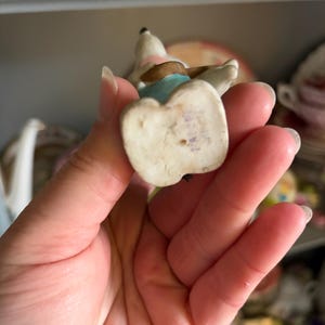 May include: A small, vintage porcelain figurine of a dog, with a white body, brown ears, and a pink and blue outfit. The figurine is held in a hand, showcasing its delicate details and aged appearance. The base of the figurine is off-white.