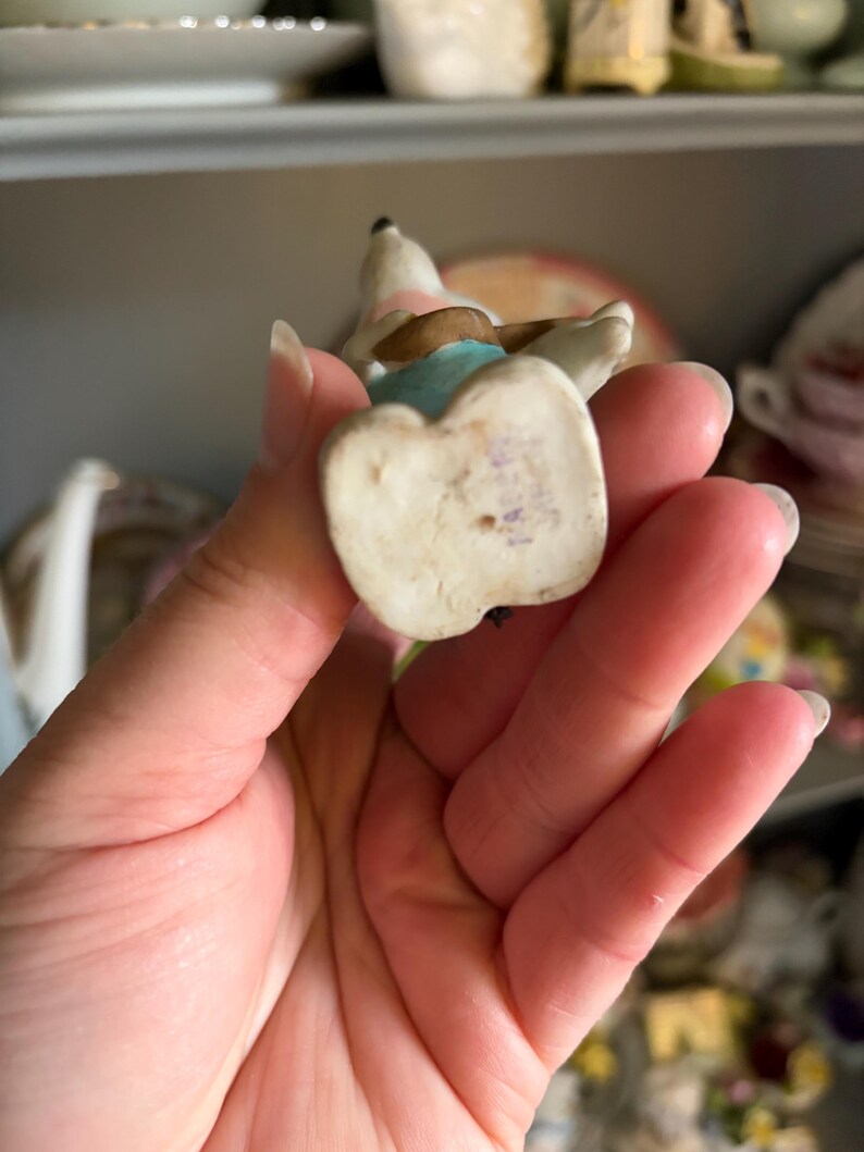 May include: A small, vintage porcelain figurine of a dog, with a white body, brown ears, and a pink and blue outfit. The figurine is held in a hand, showcasing its delicate details and aged appearance. The base of the figurine is off-white.