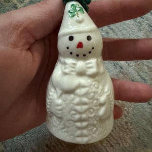 May include: A white ceramic snowman ornament with a glossy finish, featuring a green shamrock painted on the hat, a red nose, black dot eyes and mouth, a molded bow tie, and intricate embossed floral patterns on the body with a green string loop.