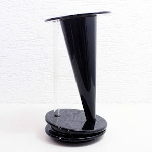 May include: A modern black vase with a flared, conical shape. The vase has a wide, flat top and sits on a stacked, oval base. A clear, cylindrical tube runs through the center, likely for holding flowers. The vase is made of a glossy material.