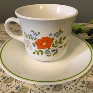 May include: A white teacup and saucer with a green rim and a floral design featuring orange, yellow, blue, and green flowers.