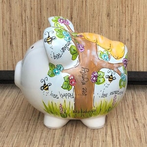 May include: White ceramic piggy bank with a painted tree and bees. The tree has the words "bee unique", "bee happy", "bee mighty", and "bee exceptional" written on it.