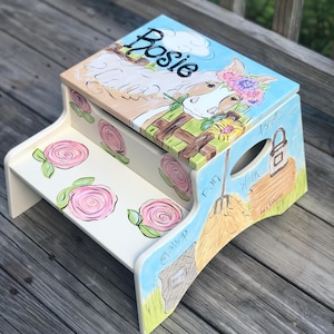 May include: A hand-painted, two-step wooden stool with a whimsical farm theme. The top features a horse with a floral crown and the name "Rosie". Pink roses adorn the steps, and the sides depict a hay bale and farm tools.