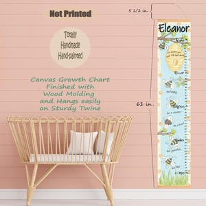 May include: A canvas growth chart with a bee theme, measuring 61 inches tall. The chart features a light blue background with illustrations of bees, a beehive, and the name "Eleanor". It is finished with wood molding and hangs easily on sturdy twine.