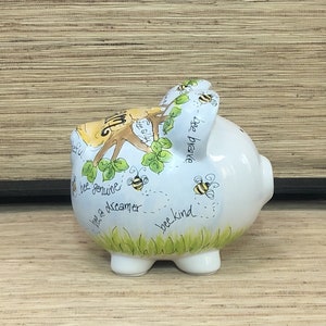 Puede incluir: Hucha de cerámica blanca con un diseño caprichoso de abejas. El banco presenta abejas pintadas a mano, hojas verdes y las frases "bee genuine", "bee brave" y "bee kind". La hucha tiene una ranura en la parte superior para insertar monedas.
