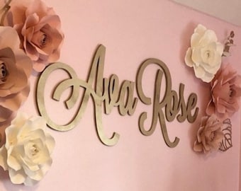 May include: A pink nursery wall decorated with a wooden sign that reads "Ava Rose" in gold script. The sign is surrounded by large paper flowers in shades of pink and white.