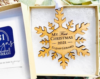 May include: A wooden snowflake ornament with the text "My First Christmas - 2021 - Elijah Gerard" engraved on it. The ornament is in a white box with a white ribbon.