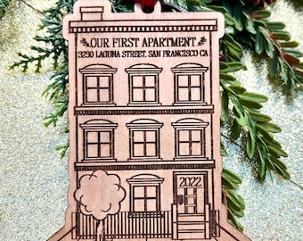 May include: A wooden ornament shaped like a house with the text "Our First Apartment" and "3250 Laguna Street San Francisco CA" on the front. The house has three windows on each of the top two floors, two windows on the bottom floor, and a door with the number "2022" above it. There is a small tree in front of the house.