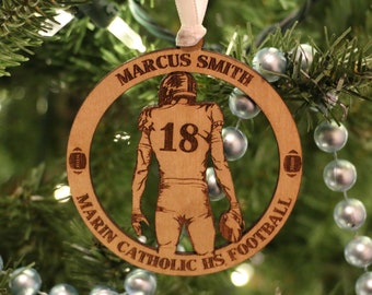 May include: A wooden Christmas ornament featuring a silhouette of a football player with the number 18 on their jersey. The ornament is personalized with the name "Marcus Smith" and the text "Marin Catholic HS Football".