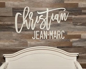 May include: White wooden crib with a white headboard and footboard. The crib has a white mattress with a gray and pink patterned sheet. The crib is in front of a wall with a wooden plank design. The wall has a white sign with the text "Christian Jean-Marc" in cursive.