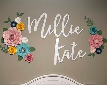 May include: A white crib with a white headboard and a floral patterned quilt. The wall behind the crib has a white wooden sign with the names "Millie Kate" written in cursive. The wall is decorated with paper flowers in shades of pink, yellow, blue, and green.