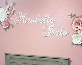 May include: A pink nursery with a crib and a wall decorated with paper flowers and the names "Mirabelle Isela" in white cursive lettering.