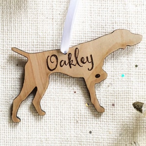 Può includere: Un ornamento natalizio in legno a forma di cane con il nome "Oakley" inciso su di esso. L'ornamento è appeso a un nastro bianco.