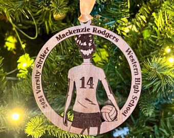 May include: Wooden Christmas ornament featuring a volleyball player with the name "Mackenzie Rodgers", the number "14", and the text "2024 Varsity Striker Western High School". The ornament is hanging from a beige ribbon.