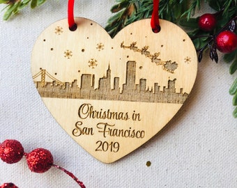 May include: A wooden heart-shaped Christmas ornament with a laser-engraved silhouette of the San Francisco skyline, snowflakes, and Santa Claus flying over the city. The ornament reads "Christmas in San Francisco 2019".