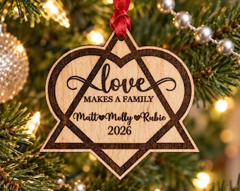 May include: A light-colored wooden ornament shaped like a heart inside a star, featuring a red ribbon loop. The text reads Love Makes A Family, followed by the names Matt, Molly, and Rubie separated by heart icons, and the year 2026 at the bottom.