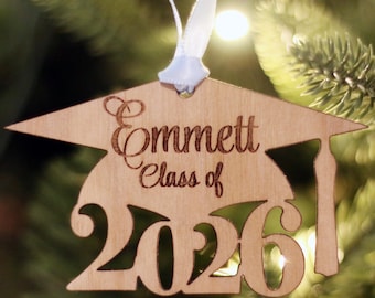 May include: Wooden graduation ornament with the name "Emmett" and "Class of 2026" engraved. The ornament is shaped like a graduation cap and has a white ribbon for hanging. The background is a blurred Christmas tree.