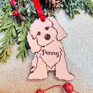 May include: A wooden dog ornament featuring the name "Penny". The light brown ornament has a red ribbon for hanging. The dog is a poodle-like breed. The background includes greenery and red berries, typical of festive decorations.
