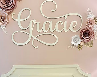 May include: A white wooden sign with the name "Gracie" written in cursive script. The sign is hanging on a pink wall with a bed in the foreground. The wall is decorated with paper flowers in shades of pink and white.