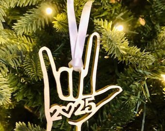 May include: A wooden Christmas ornament in the shape of a hand making the "I love you" sign. The ornament has the year "2025" with two hearts cut out. It hangs from a white ribbon against a backdrop of a Christmas tree.