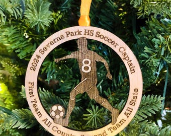 May include: Wooden soccer ornament with a player silhouette, number 8, and the text "2024 Severna Park HS Soccer Captain." Includes "First Team All County, Second Team All State." The ornament is hanging on a Christmas tree.