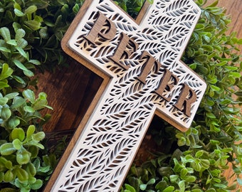 May include: A white wooden cross with a leafy pattern and the name "Peter" carved into the wood. The cross is surrounded by green foliage.