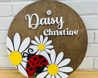 May include: A round wooden sign with the names "Daisy Christine" in white lettering. The sign is decorated with white daisies and a red ladybug.