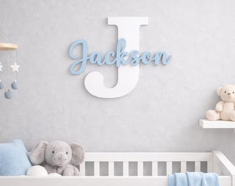 May include: A white wooden letter "J" with the name "Jackson" in light blue script, mounted on a gray wall. The nursery decor includes a crib, stuffed animals, and a mobile.