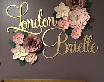 May include: Gold cursive lettering spells "London Brielle" above a crib. The names are surrounded by paper flowers in shades of pink, cream, and brown, with green leaf accents. The wall is a muted purple.