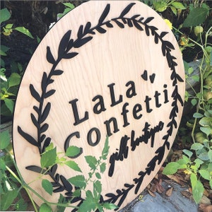 Puede incluir: Un letrero redondo de madera con un dise&ntilde;o de corona negra y el texto "LaLa Confetti gift boutique".